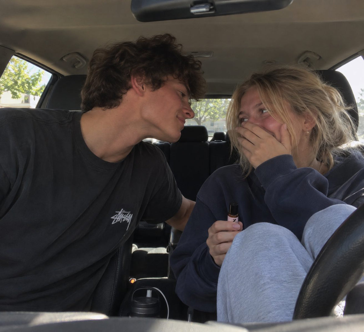 a couple in a car, her hand over her mouth laughing, holding the sirense bottle